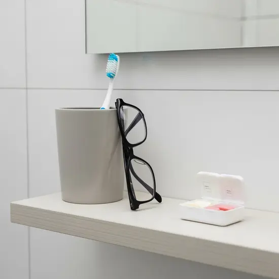 Symbolbild zu Zusatzversicherungen: Zahnbürste in Becher neben Brille und kleiner Medikamentenbox auf sauberem Badregal.