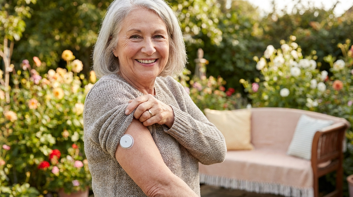 Seniorin zeigt lächelnd einen kleinen runden Glukosesensor an ihrem Oberarm.