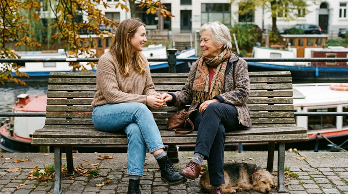 Eine jüngere Frau spricht auf Augenhöhe mit einer älteren Dame, beide lächeln sich freundlich an, entspannte Körperhaltung.