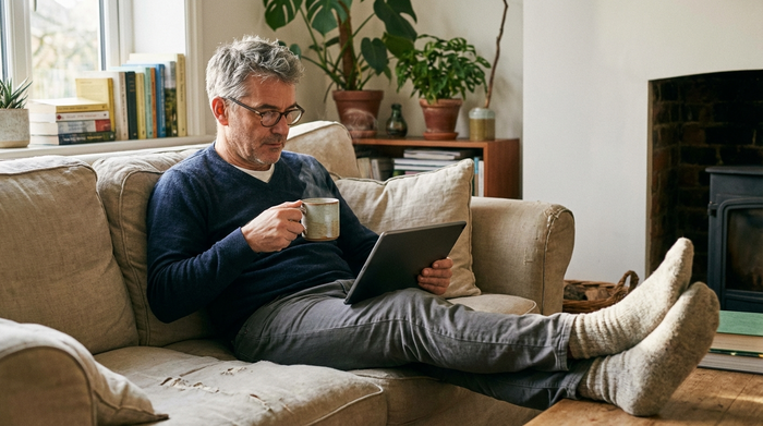 Ein Mann mittleren Alters sitzt entspannt mit einer Tasse Tee auf dem Sofa und schaut sich konzentriert ein Lernvideo auf dem Tablet an.