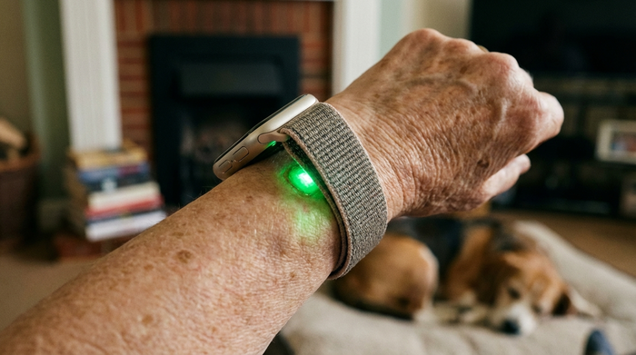 Nahaufnahme des Handgelenks einer Seniorin, an dem eine Smartwatch mit leuchtend grünem Sensor auf der Rückseite anliegt, der sanft die Haut beleuchtet. Im Hintergrund unscharf ein gemütliches Wohnzimmer.