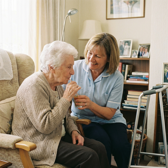 Pflegerin hilft einer Seniorin behutsam beim Trinken aus einem Glas
