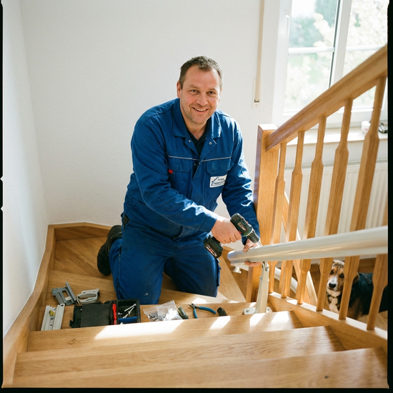 Freundlicher Monteur in Arbeitskleidung installiert sorgfältig die Schiene eines Treppenlifts auf einer hellen Holztreppe