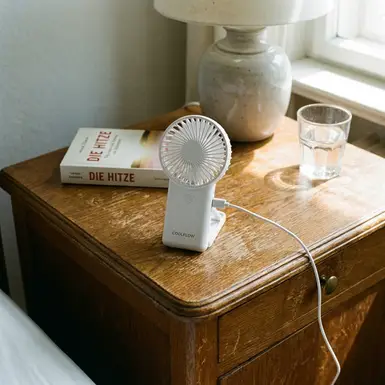 Kleiner weißer Handventilator steht einsatzbereit auf einem Holz-Nachttisch