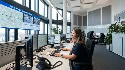 Eine aufmerksame Mitarbeiterin sitzt mit einem Headset an ihrem Arbeitsplatz in einer modernen Notrufzentrale.