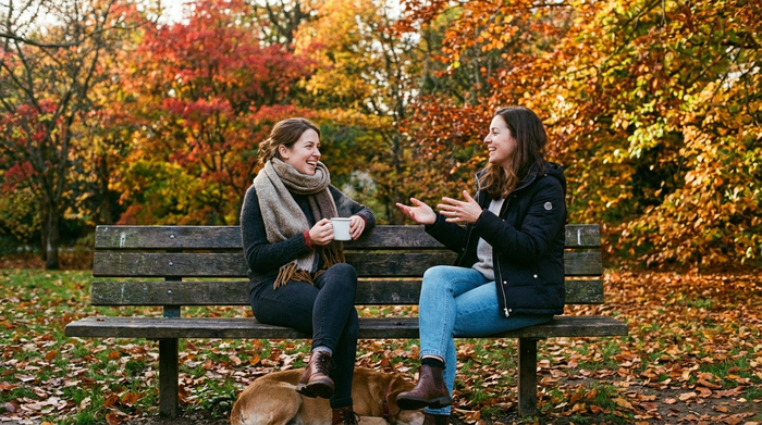 Zwei Frauen sitzen entspannt auf einer Parkbank und unterhalten sich, herbstliche Bäume im Hintergrund