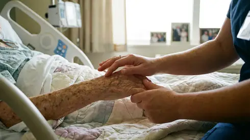 Sanfte und liebevolle Berührung der Hand eines älteren Menschen im Pflegebett durch eine Pflegekraft