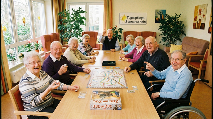 Senioren sitzen fröhlich gemeinsam an einem großen Tisch in einer Tagespflege-Einrichtung und spielen ein Gesellschaftsspiel