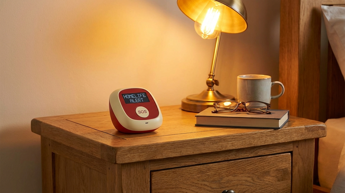 Kompaktes Hausnotruf-Gerät auf einem Nachttisch aus Holz neben einer warmen Leselampe.