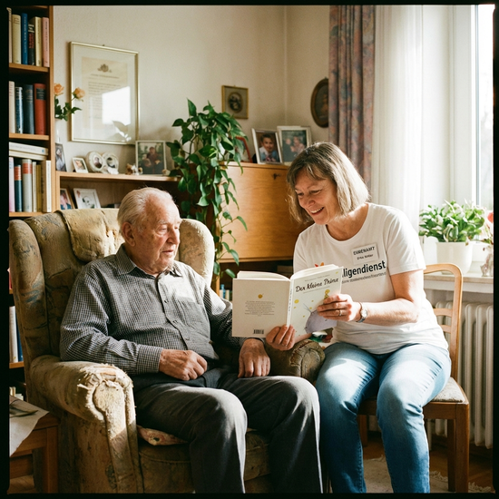 Ehrenamtliche Begleiterin beim Vorlesen aus einem Buch für einen älteren Herrn im Sessel