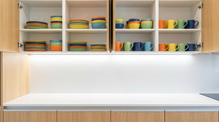 Geöffnete Küchenschränke mit ordentlich aufgereihten, bunten Tellern und Tassen. Die Arbeitsfläche darunter ist komplett leer und aufgeräumt, hell und blendfrei beleuchtet durch eine moderne LED-Leiste.
