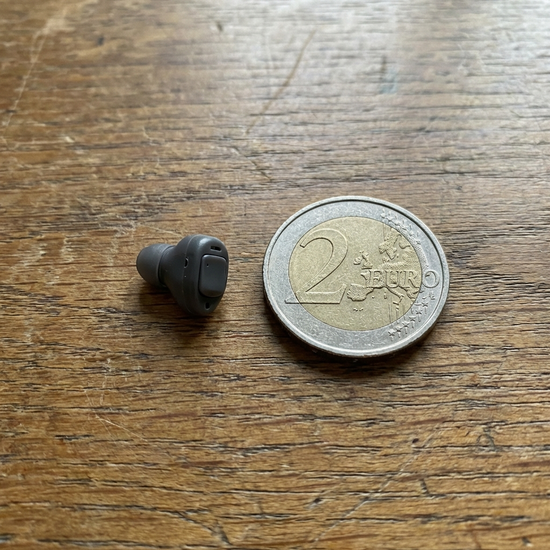 Modernes, unauffälliges Hörgerät liegt neben einer 2-Euro-Münze zum Größenvergleich auf einem Holztisch