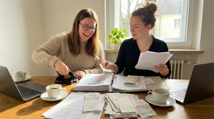 Zwei Frauen sitzen am Tisch und sortieren Quittungen und Fahrkarten in einen Ordner.
