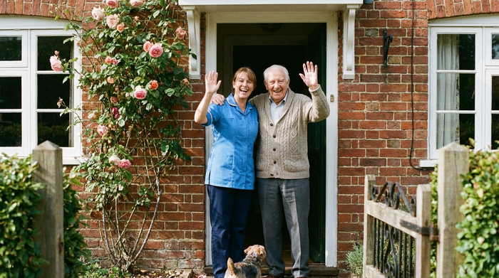 Eine Pflegekraft und ein Senior stehen lachend an der Haustür und winken.