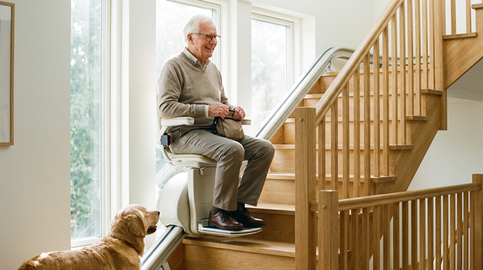 Freundlicher Senior fährt entspannt auf einem modernen Sitzlift eine helle Holztreppe hinauf