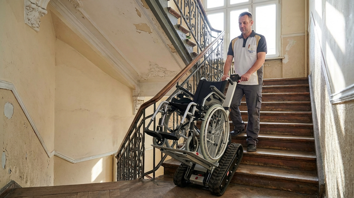 Ein professioneller Pfleger bedient eine moderne Treppenraupe, mit der ein leerer Rollstuhl sicher die Stufen eines typischen Berliner Altbaus hinauf transportiert wird.