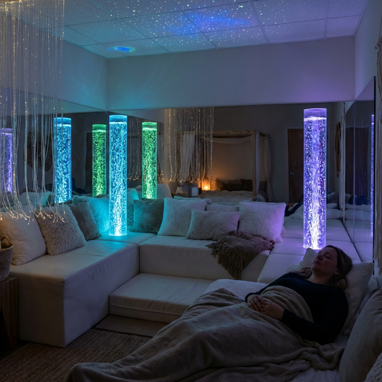 Ein gemütlich eingerichteter Snoezelen-Raum mit gedimmtem, sanftem Licht. Bequeme weiße Liegelandschaften, weiche Kissen und sanft blubbernde, farbig beleuchtete Wassersäulen schaffen eine beruhigende Atmosphäre für tiefenentspannte Momente.