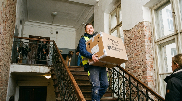 Ein freundlicher Lieferant in Arbeitskleidung trägt vorsichtig ein gut verpacktes medizinisches Gerät durch das Treppenhaus eines typischen Hamburger Mehrfamilienhauses.