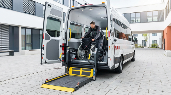 Ein moderner Kleinbus mit ausgefahrener, stabiler hydraulischer Hebebühne am Heck. Ein Fahrer sichert routiniert einen leeren Rollstuhl im hellen Innenraum des Fahrzeugs. Fokus auf Sicherheit und Technik, saubere Umgebung.