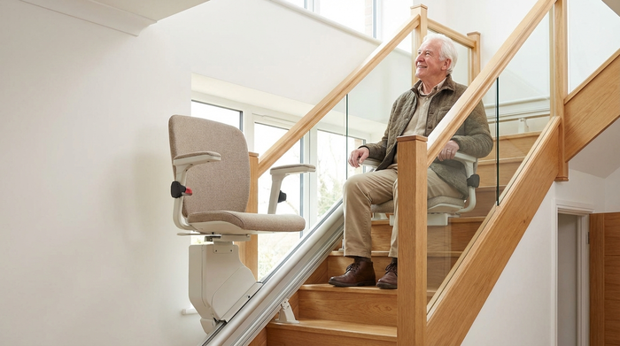 Ein moderner, unauffälliger Treppenlift in einem hellen, gepflegten Treppenhaus aus Holz. Ein lächelnder Senior sitzt bequem und sicher auf dem Sitz, während er nach oben fährt. Realistische Architektur, saubere Umgebung.