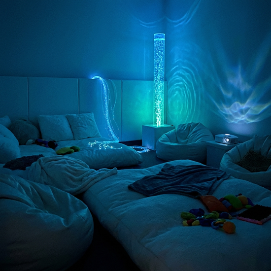 Ein gemütlicher Snoezelen-Raum mit gedimmtem, beruhigendem bläulichem Licht. Bequeme weiße Liegelandschaften, eine sanft blubbernde Wassersäule im Hintergrund. Friedliche und entspannende Atmosphäre, realistisch.