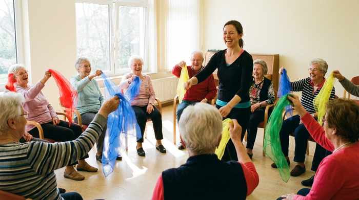 Eine Gruppe von fröhlichen Senioren sitzt in einem hellen Raum im Kreis zusammen und macht leichte Gymnastikübungen mit bunten Tüchern. Eine motivierende Betreuerin leitet die Gruppe an. Lebhafte, positive und aktive Stimmung.