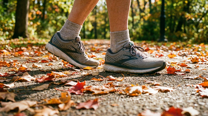 Nahaufnahme der Füße einer älteren Person in bequemen, stützenden Laufschuhen auf einem gepflegten Parkweg, umgeben von herbstlichen Blättern. Fokus auf die stabile und gesunde Fußhaltung beim Gehen.