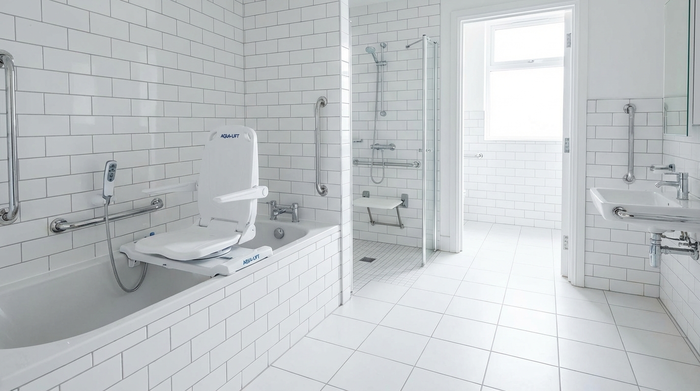 Ein sauberes, barrierefreies Badezimmer mit weißen Fliesen. In der Badewanne ist ein moderner, weißer Badewannenlift montiert, bereit zur sicheren Nutzung.