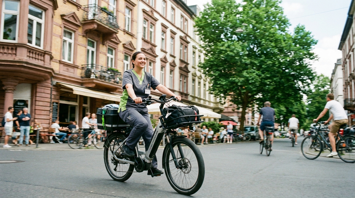 Eine moderne Pflegekraft fährt mit einem E-Bike durch eine belebte Straße im Frankfurter Nordend. Im Hintergrund sind unscharf typische Altbaufassaden und grüne Bäume zu erkennen. Helles Tageslicht, dynamische und positive Stimmung.