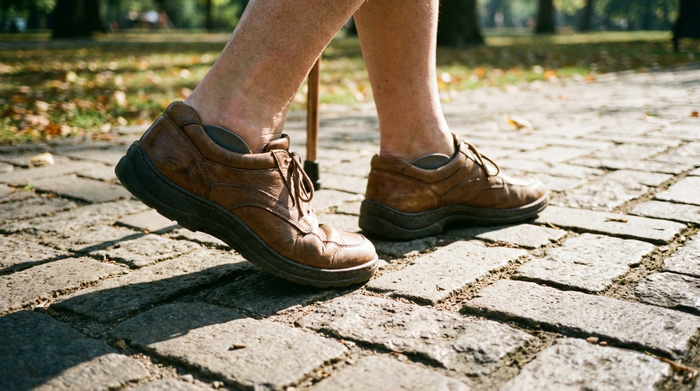 Nahaufnahme der Füße eines älteren Menschen in bequemen, stützenden Lederschuhen beim Spaziergang auf einem gepflasterten Weg im Park. Realistisch, helles Tageslicht, Fokus auf gesundes Gehen.