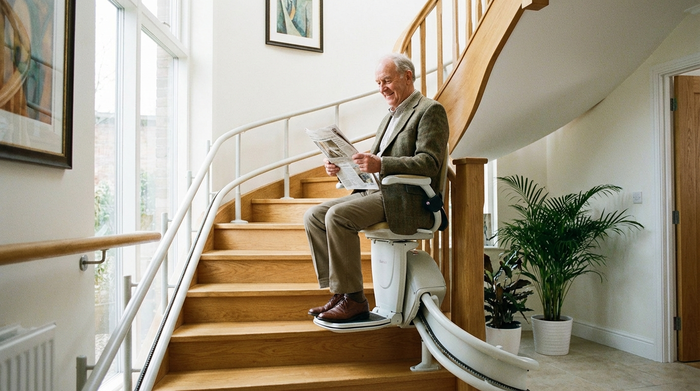 Ein älterer Mann fährt sicher und entspannt auf einem modernen Treppenlift eine geschwungene Holztreppe hinauf. Helles Treppenhaus, gepflegte Umgebung, Fokus auf Mobilität und Sicherheit.