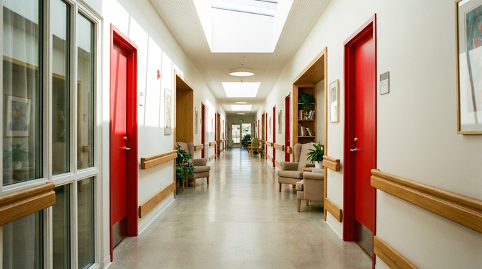 Ein heller, endloser Flur in einer modernen Pflegeeinrichtung mit farblich abgesetzten, roten Türen und Handläufen aus Holz. Tageslicht durchflutet den Gang, gemütliche Sitznischen laden zum Verweilen ein.