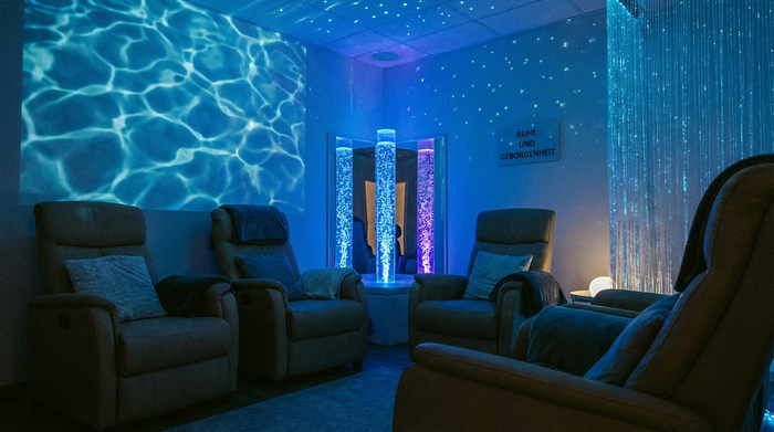Ein gemütlicher Snoezelen-Raum mit gedimmtem, bläulichem Licht, bequemen Liegesesseln und sanft leuchtenden Wassersäulen. Eine entspannte Atmosphäre, die tiefe Ruhe und Geborgenheit ausstrahlt.