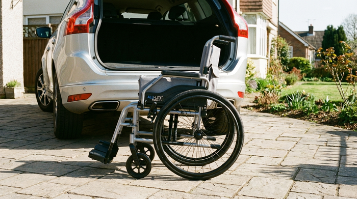 Ein moderner, leichter Aluminium-Rollstuhl steht ordentlich zusammengeklappt neben dem geöffneten Kofferraum eines Autos auf einer gepflasterten Auffahrt an einem sonnigen Tag.