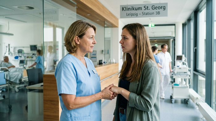 Eine professionelle Pflegekraft in hellblauer Dienstkleidung spricht auf dem Flur eines modernen Leipziger Krankenhauses einfühlsam mit einer besorgten Tochter. Im Hintergrund verschwommene Krankenhausumgebung, helles Tageslicht.