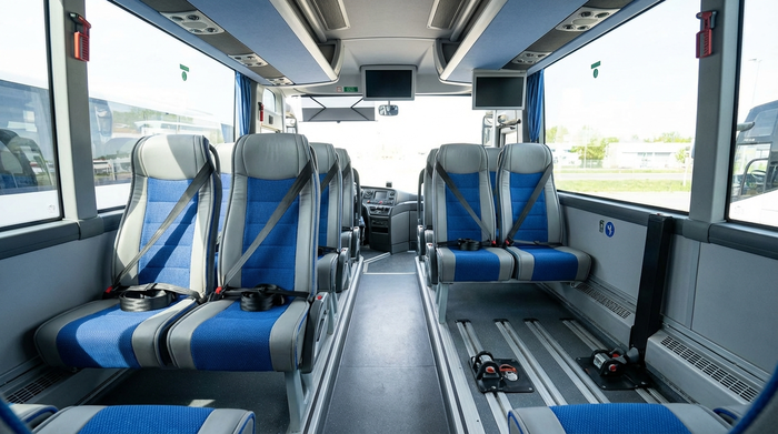 Das aufgeräumte Innere eines modernen Transportbusses. Bequeme, saubere Sitze mit sichtbaren Sicherheitsgurten und speziellen Halterungen am Boden. Helles Tageslicht fällt durch die großen Fenster, sehr realistisch.