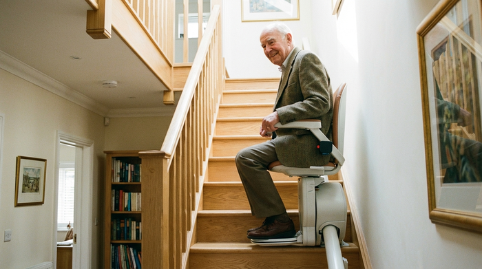 Ein älterer Herr fährt lächelnd auf einem modernen Treppenlift eine helle Holztreppe hinab. Er wirkt sicher und zufrieden. Gepflegtes Treppenhaus, realistische, hochwertige Fotografie ohne Text.