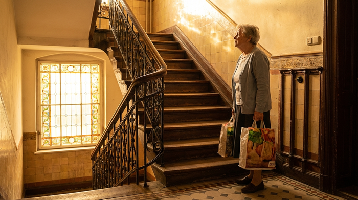 Eine ältere Frau steht mit Einkaufstaschen vor einer steilen Holztreppe in einem klassischen Leipziger Gründerzeithaus, das warme Sonnenlicht fällt durch das Treppenhausfenster.
