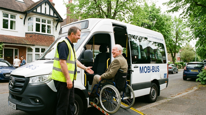 Ein rollstuhlgerechter Kleinbus holt einen fröhlichen älteren Herrn an seiner Haustür ab, der Fahrer hilft ihm freundlich beim Einstieg. Helles Tageslicht, gepflegte Wohngegend, realistische Szene.