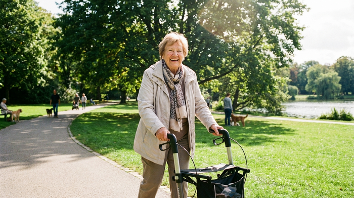 Eine glückliche ältere Frau spaziert mit einem modernen Rollator durch einen sonnigen, grünen Park. Sie trägt eine leichte Übergangsjacke und lächelt zufrieden. Im Hintergrund große Bäume und ein gepflasterter Weg.