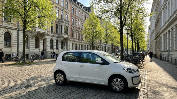Ein kleines, weißes Elektroauto parkt in einer typischen Straße in Hannover mit schönen Altbauten und grünen Bäumen. Frühlingswetter, sonnig, klare Szene ohne lesbare Logos oder Text.