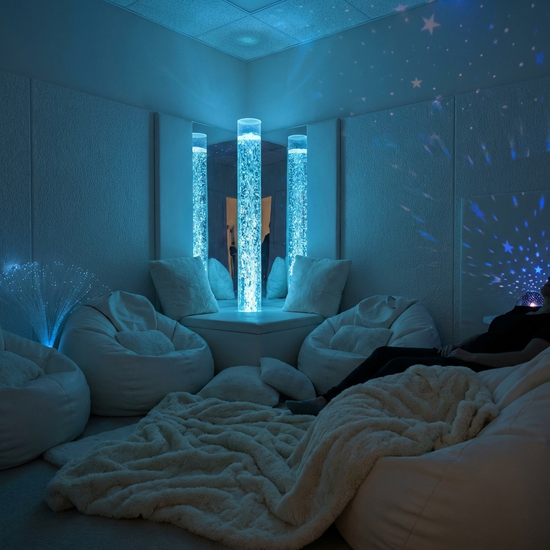 Ein gemütlicher Snoezelen-Raum mit gedimmtem, beruhigendem blauem Licht. Eine leuchtende Wassersäule mit sanften Blasen steht in der Ecke, umgeben von weichen weißen Sitzkissen und flauschigen Decken. Entspannende und therapeutische Atmosphäre, ohne Text.