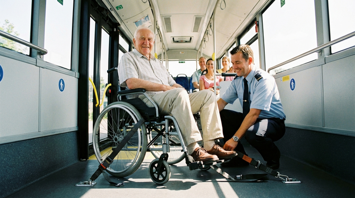 Innenansicht eines geräumigen Transportbusses. Ein älterer Herr sitzt sicher und entspannt in einem Rollstuhl, der fachgerecht mit Gurten am Boden verankert ist. Ein Fahrer prüft lächelnd den Sitz der Sicherheitsgurte. Realistisch, hell und freundlich.