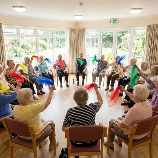 Senioren sitzen im Kreis in einem hellen Raum und machen leichte Sitzgymnastik mit bunten Tüchern. Alle wirken konzentriert und haben Freude an der Bewegung.