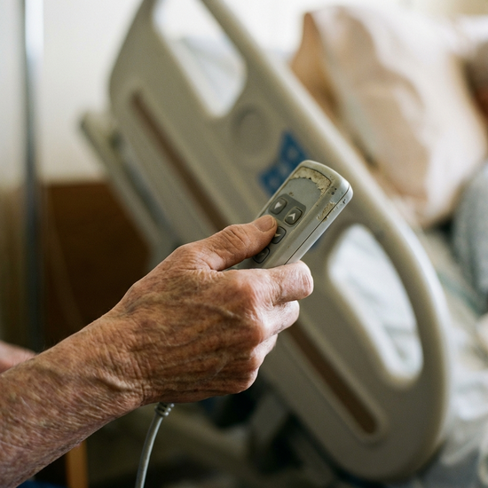 Ein Detailbild eines elektrischen Handschalters für ein Pflegebett, gehalten von einer älteren Hand. Im unscharfen Hintergrund das hochgefahrene Kopfteil eines Bettes. Realistische Beleuchtung, ohne Text.