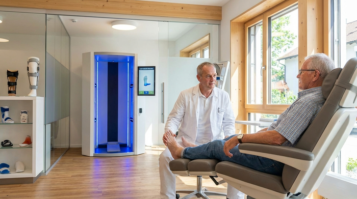 Ein moderner, heller Raum in einem orthopädischen Fachgeschäft. Ein erfahrener Orthopädietechniker in weißem Kittel untersucht behutsam den Fuß eines älteren Herrn, der entspannt auf einem Behandlungsstuhl sitzt. Im Hintergrund ist ein leuchtendes, modernes 3D-Scan-Gerät zu erkennen.