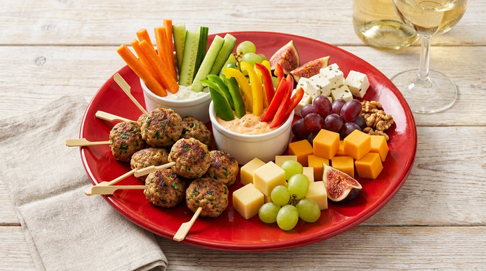 Verschiedene bunte, appetitlich angerichtete Fingerfood-Snacks wie kleine Frikadellen, Gemüsesticks und Käsewürfel auf einem leuchtend roten Teller, der auf einem hellen Holztisch steht.
