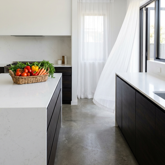 Eine saubere, moderne Küche mit glänzenden Arbeitsflächen. Ein Korb mit frischem Gemüse steht auf dem Tresen, im Hintergrund weht eine weiße Gardine.