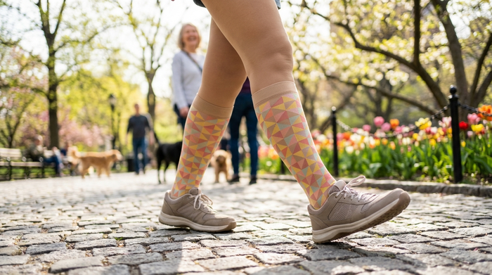 Die Beine einer aktiven Seniorin, die modische, blickdichte Kompressionsstrümpfe trägt und in bequemen Schuhen auf einem gepflasterten Weg in einem Park spaziert. Frühlingshaftes Wetter, fröhliche Stimmung, Fokus auf die Beine und Schuhe.