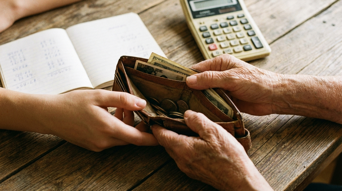 Zwei Hände, eine jung und eine alt, halten gemeinsam eine geöffnete Geldbörse auf einem Holztisch. Im Hintergrund sind unscharf ein Notizbuch und ein Taschenrechner zu erkennen.